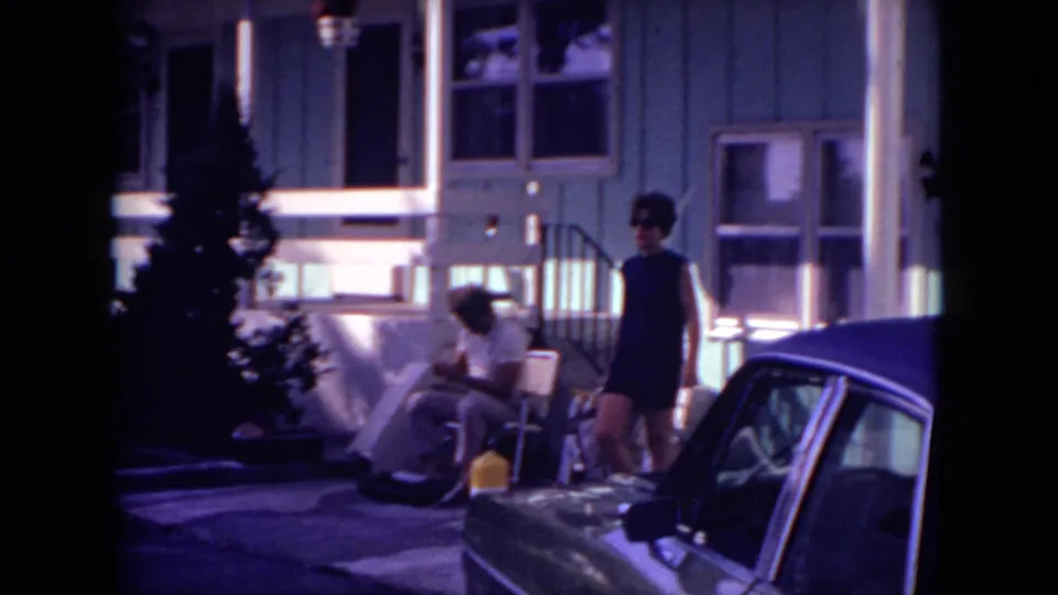 1971 Mississippi residential scene with a woman standing beside a classic car, porch furniture, front steps, and a seated person in bright sunlight.