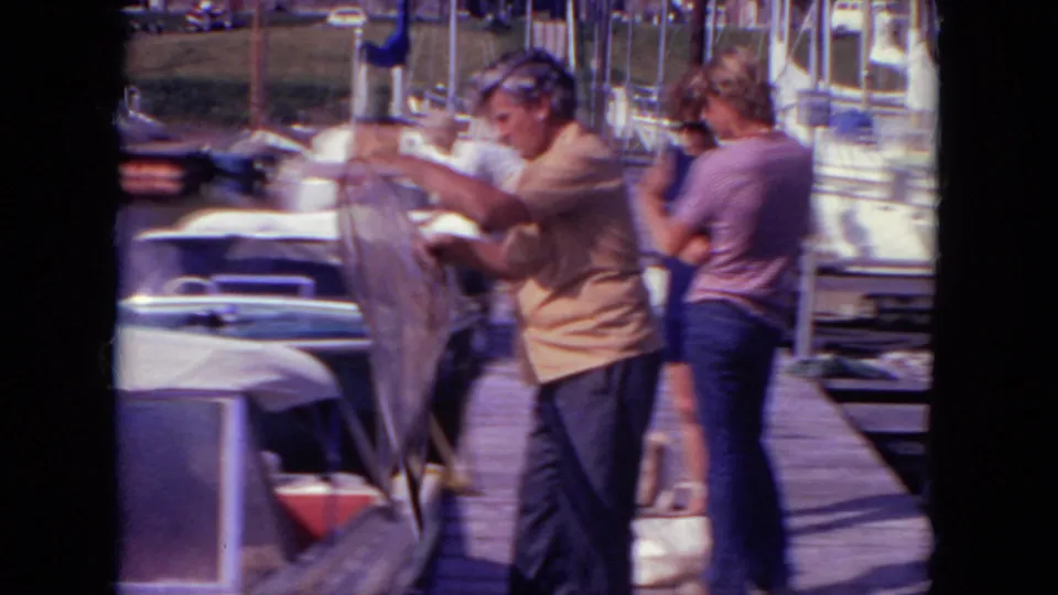 1971 Mississippi dock scene with adults in casual clothing near small boats, one person lifting or arranging fabric while another stands nearby.