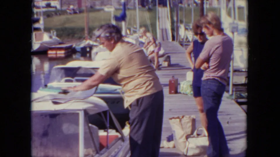 1971 Mississippi home-movie frame showing a man in a yellow shirt working near boats on a dock, with other adults and bags nearby.