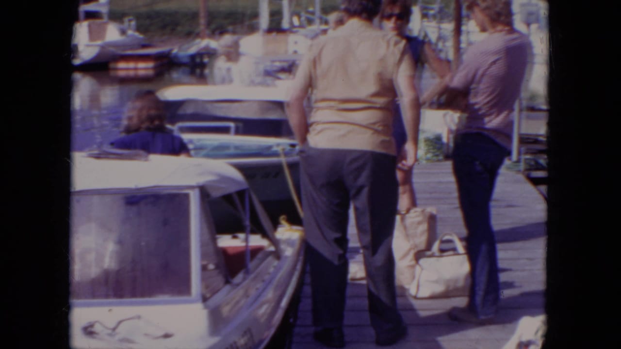 1971 Mississippi home-movie frame showing adults on a wooden dock near moored motorboats under bright sun.