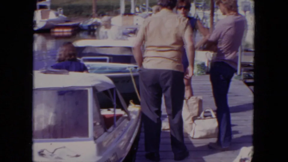1971 Mississippi home-movie frame showing adults on a wooden dock near moored motorboats under bright sun.