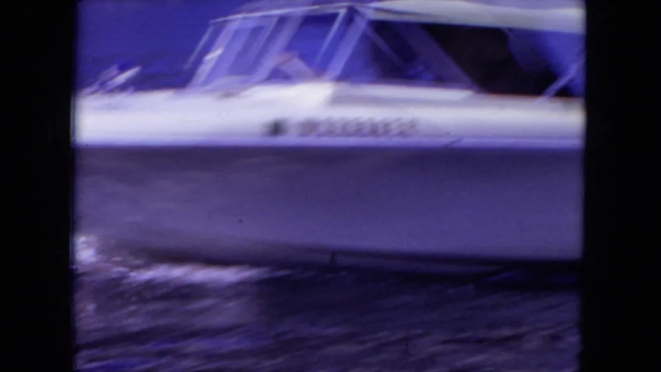 A white speed boat crosses blue Mississippi lake water in a grainy 8mm frame, with windows and side markings partly visible.