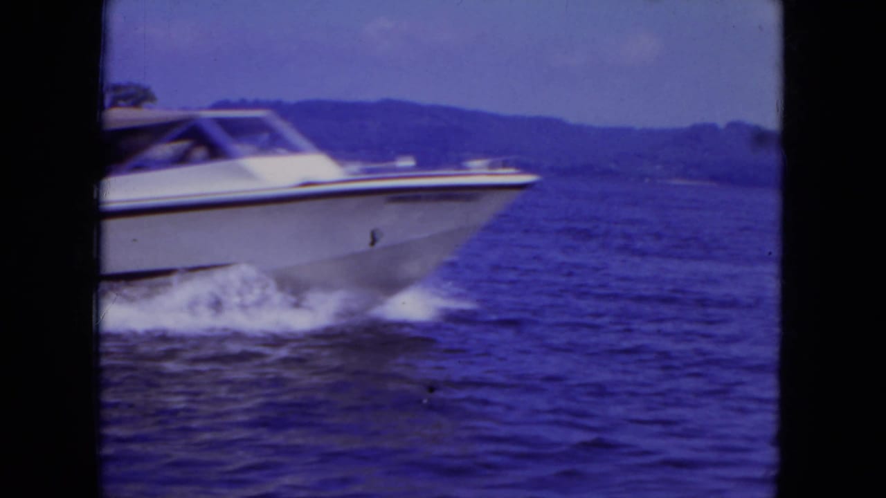White speedboat seen closer in the 1971 Mississippi home-movie footage, with rippled water and low shoreline behind it.