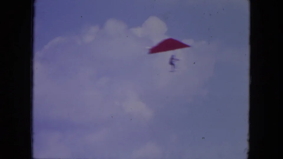 A lone hang glider with a red wing and suspended pilot silhouette drifts through a cloudy blue sky in Mississippi 1971 footage.