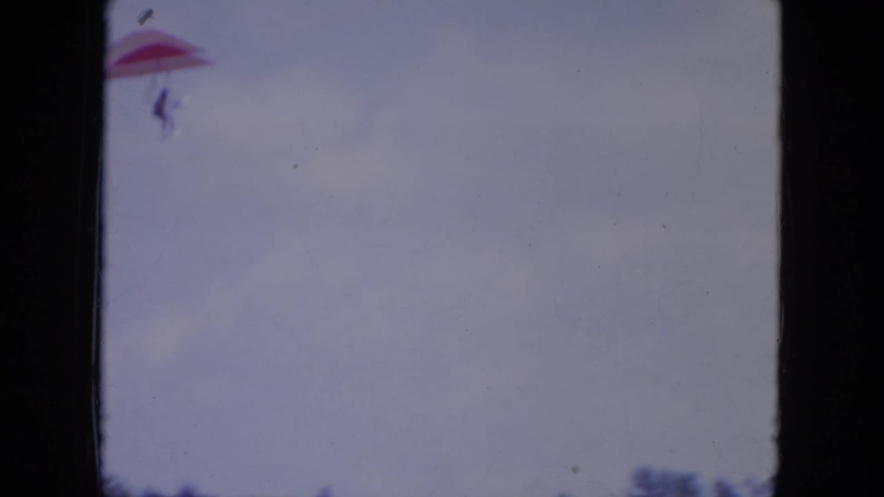 Distant red hang glider against a pale sky with dark film borders.