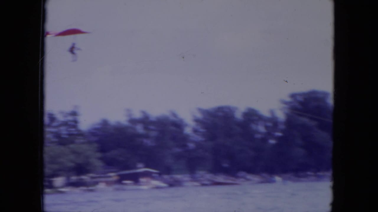 The red hang glider moves over a Mississippi lake toward a distant tree-lined shore in 1971 home-movie footage.