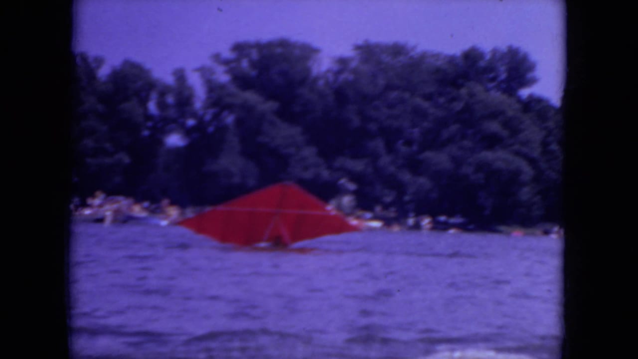 1971 Mississippi 8mm frame showing a red parasail low on the lake surface with a tree-lined shore and distant swimmers behind it.