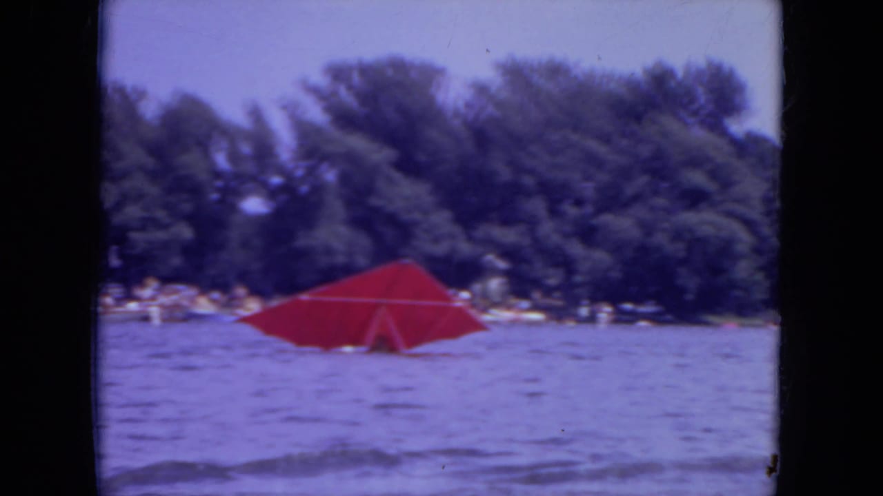 Red parasail on rippled lake water with a wooded shoreline in the distance.