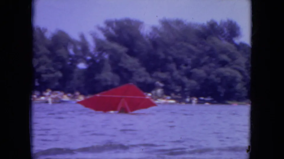 The red parasail remains on the lake in this 1971 Mississippi home-movie frame, backed by trees, distant people, and small shoreline activity.