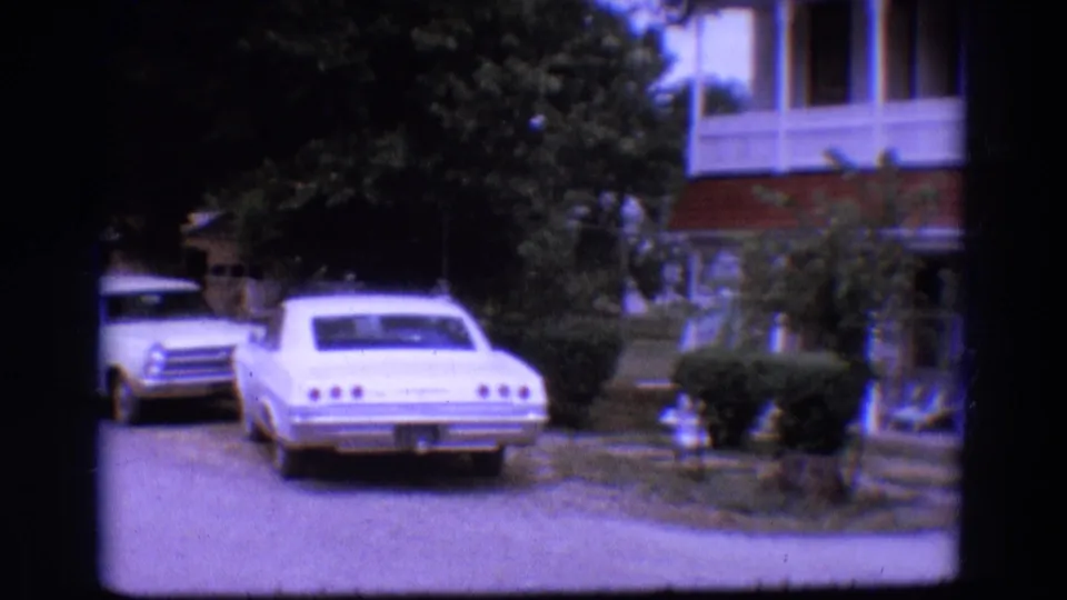 1968 Diller, Nebraska home-movie frame showing parked cars near a white two-story clapboard house and shaded driveway.