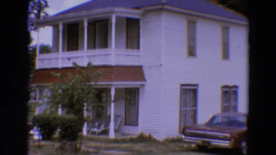 1968 Diller, Nebraska frame of a white two-story house with porch, front yard plantings, and a dark parked car partly in view.