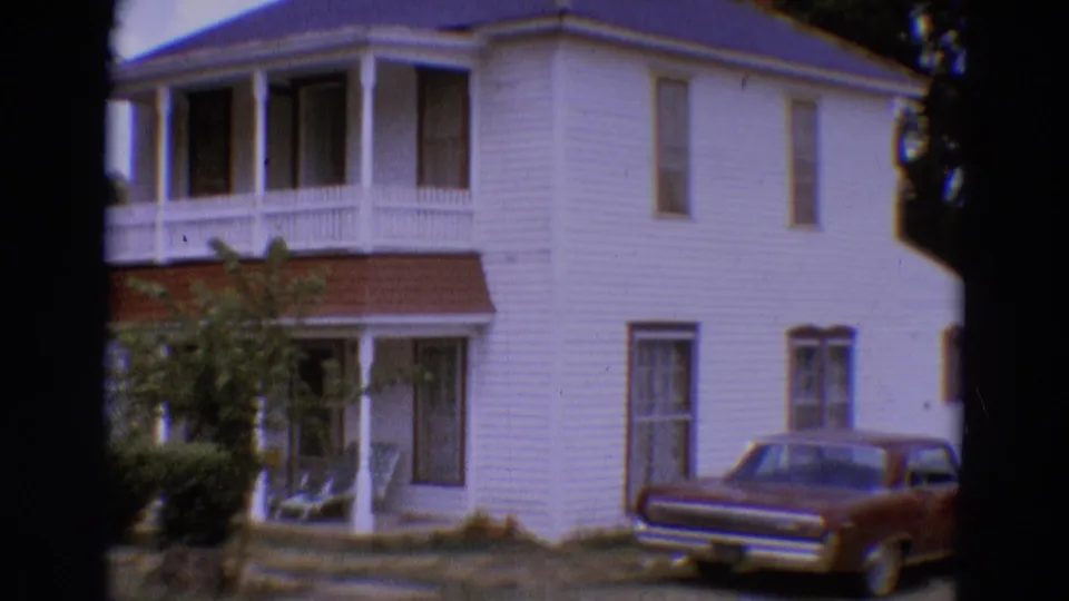 1968 Diller, Nebraska home-movie frame showing a white clapboard house, porch, trees, and a maroon sedan parked nearby.