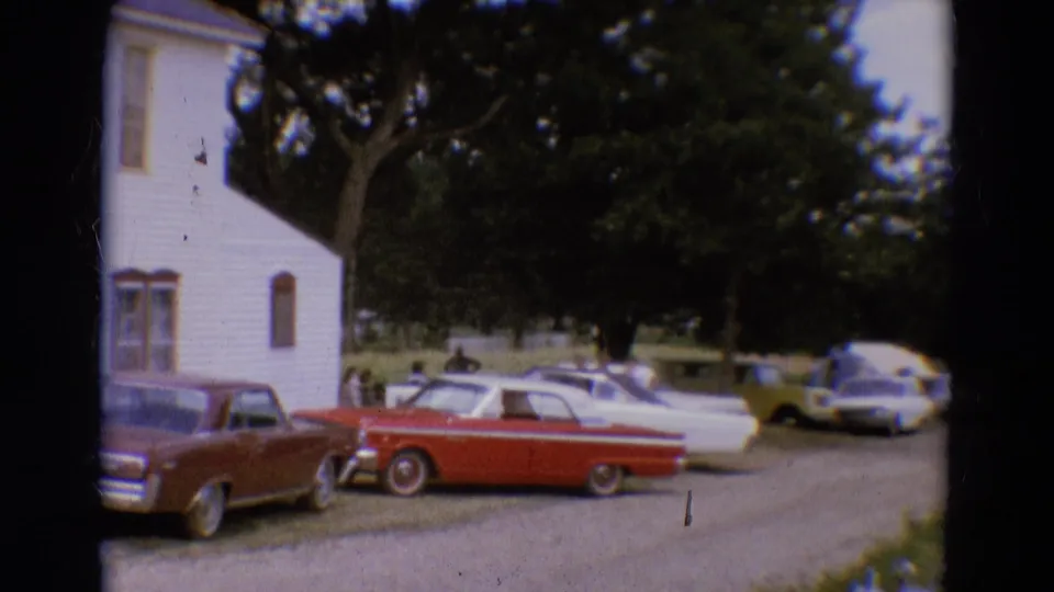 1968 Diller, Nebraska home-movie frame showing classic cars parked near a white clapboard building under leafy trees.