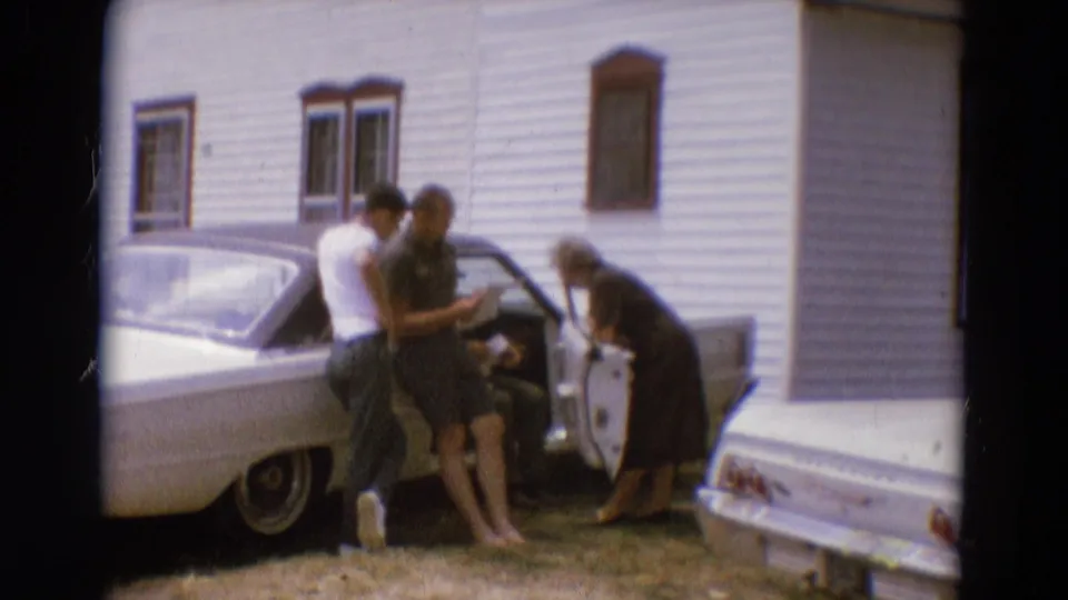 1968 Diller, Nebraska home-movie frame showing adults beside an open car door near a white clapboard building.