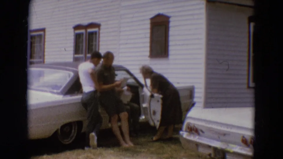 1968 Diller, Nebraska frame with adults gathered by a parked car and open door outside a clapboard building.