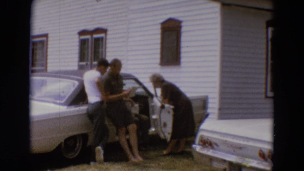 Three adults gather around an open car door beside a white-sided house.