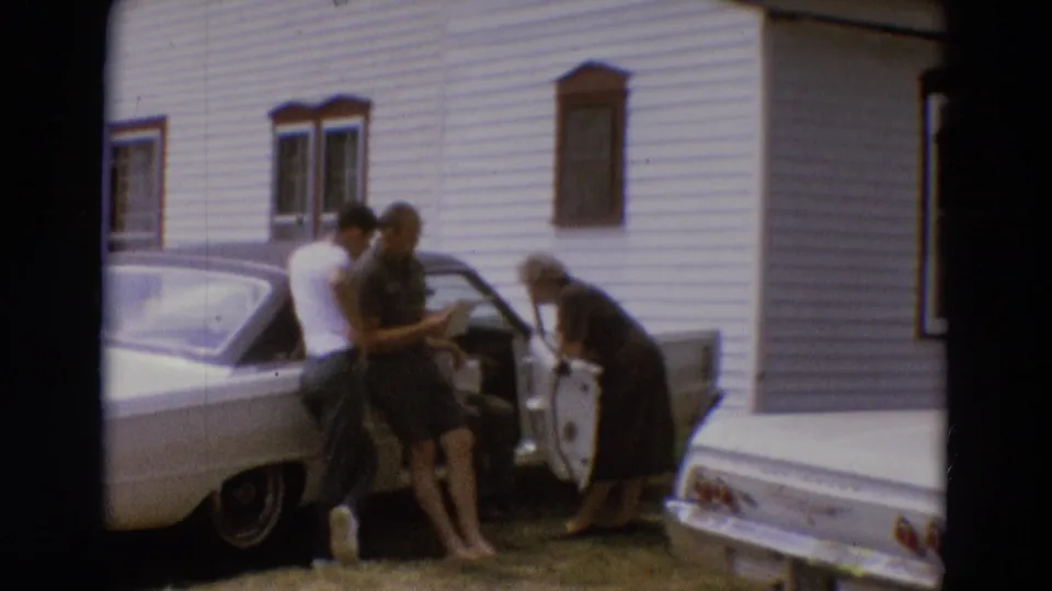 In Diller, Nebraska in 1968, three adults stand by a white car as one woman leans into the open door near a white-sided house.