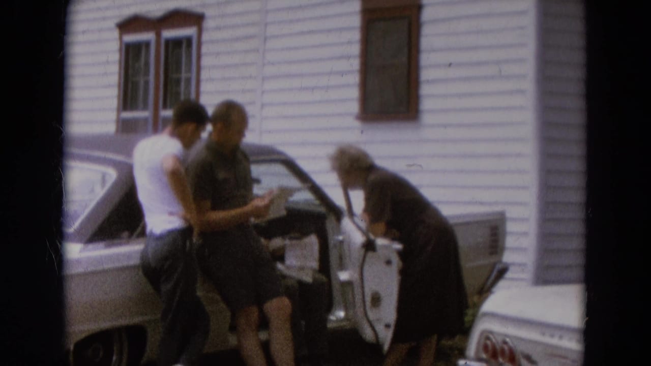 Two men look at a paper while an older woman leans into the open car door.
