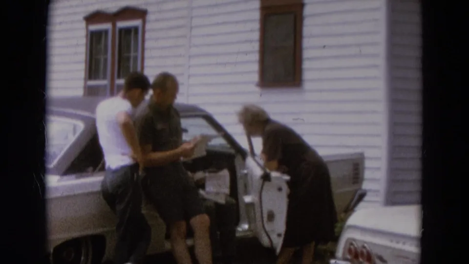 Two men in casual clothing read or examine a paper beside a white car while an older woman bends toward the open door.