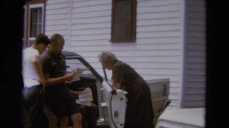 Three adults remain beside the white car in the 1968 Nebraska home-movie scene, with the paper visible between the two men.