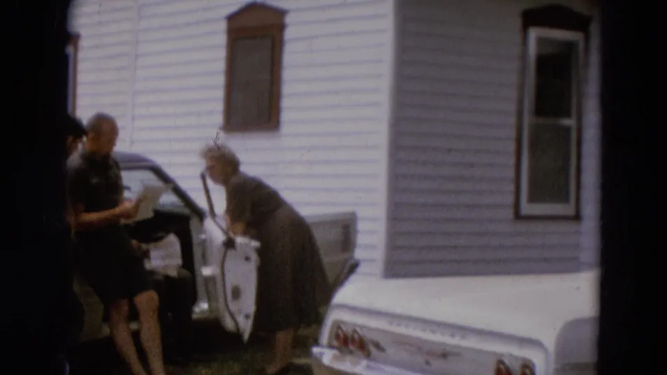 The group shifts slightly near the open white car door, with the older woman bent forward and the house exterior filling the background.