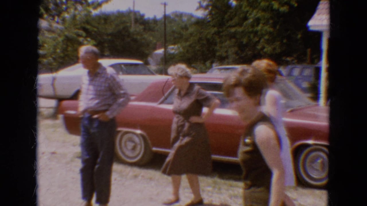 Adults stand and walk near a red sedan on a sunny gravel driveway.