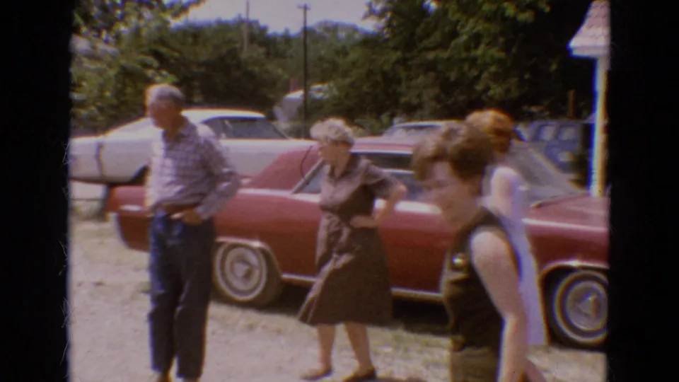 In 1968 Diller, Nebraska home-movie footage, adults gather beside a red sedan and pale parked cars on a sunny residential driveway.