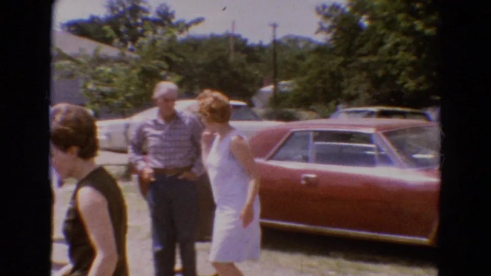 Adults in 1960s summer clothing walk and talk near a red sedan in the Diller, Nebraska residential gathering scene.
