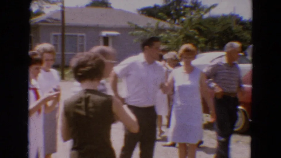 A home-movie frame shows adults conversing near parked cars, with a modest house and trees behind them in Diller, Nebraska.