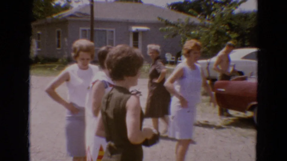 Adults in summer clothing stand and talk near a red car outside a house in the 1968 Diller, Nebraska family gathering footage.