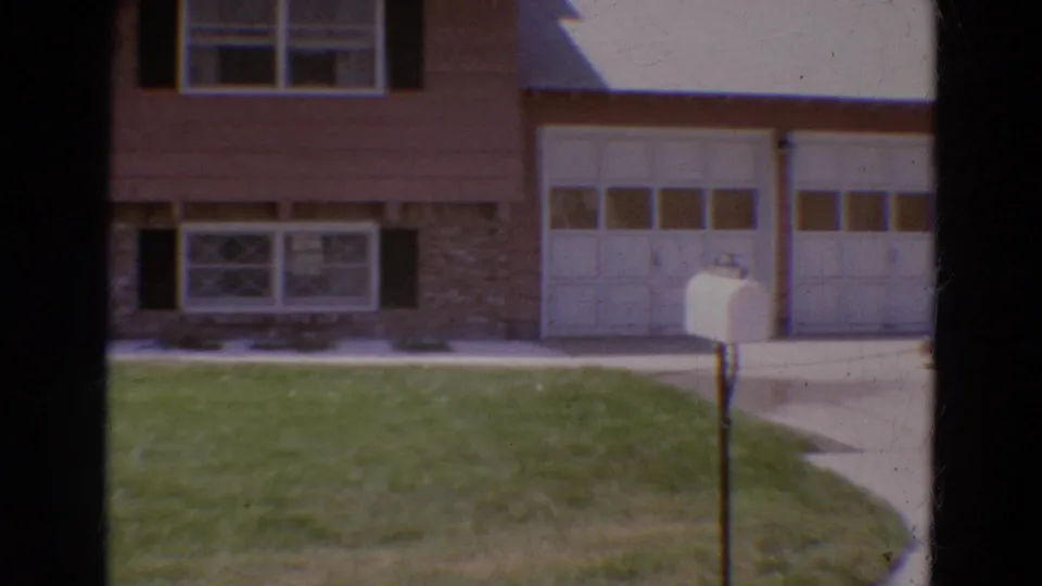 1968 Diller, Nebraska home-movie frame centered on the lower house facade, white garage doors, stonework, lawn, and mailbox.