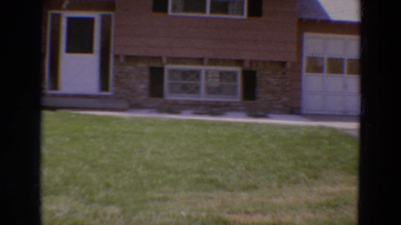 Brown house exterior with green lawn, white front door, windows, shutters, and part of a garage.