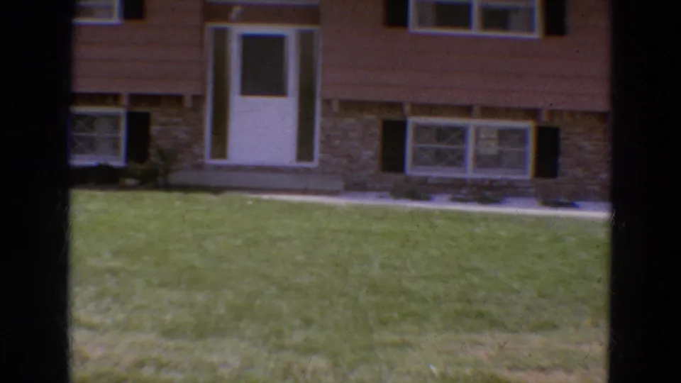 1968 Diller, Nebraska frame with the camera panning across the house front, showing lawn, white entry door, windows, shutters, and stone facade.
