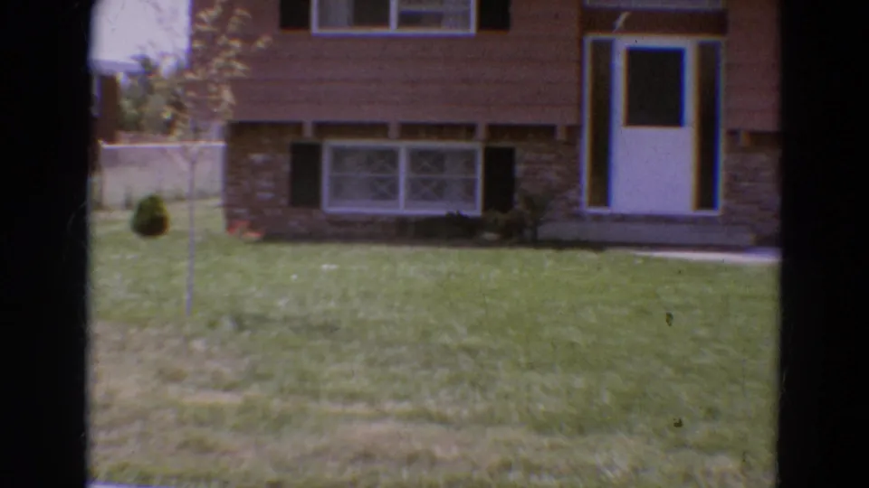 1968 Diller, Nebraska frame showing the house front from the lawn, with brown siding, stone base, white door, windows, shrubs, and yard detail.