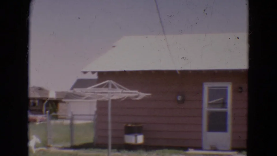 1968 Diller, Nebraska frame showing the side of a pink house with a back door, window, fence, drying rack, and small dog in the yard.