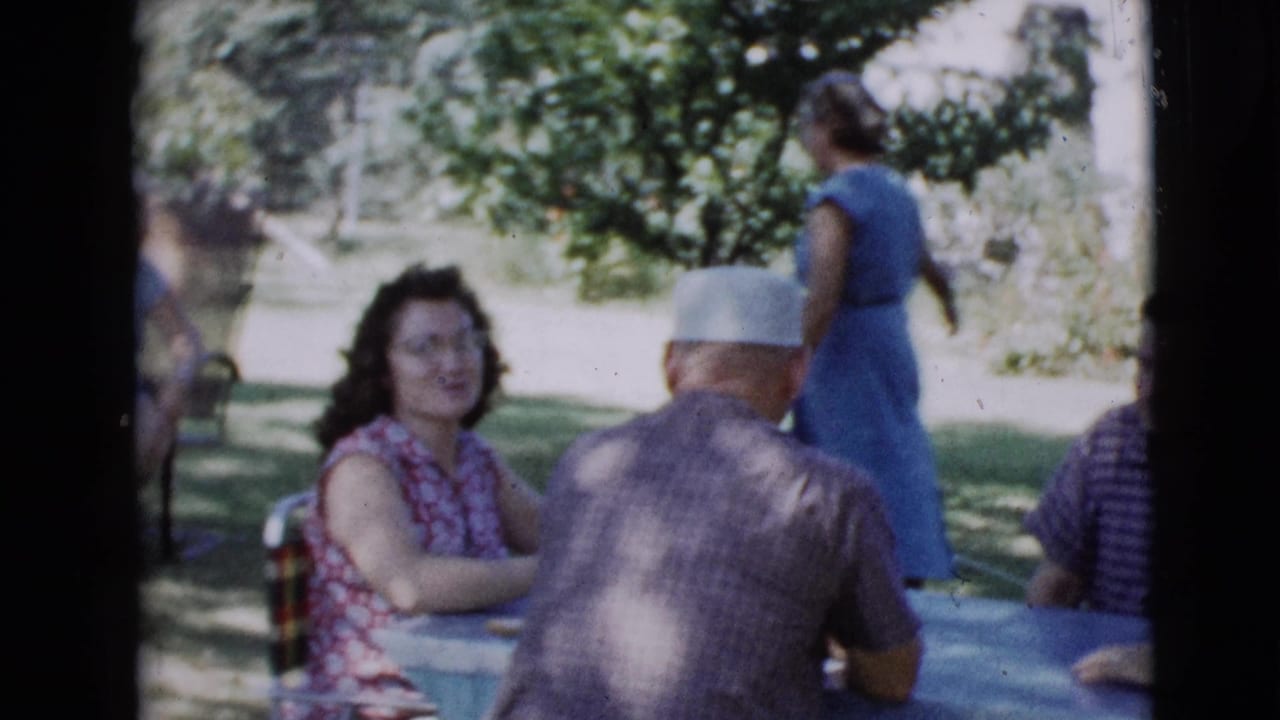Adults sit at an outdoor table while a woman in a blue dress walks in the background.