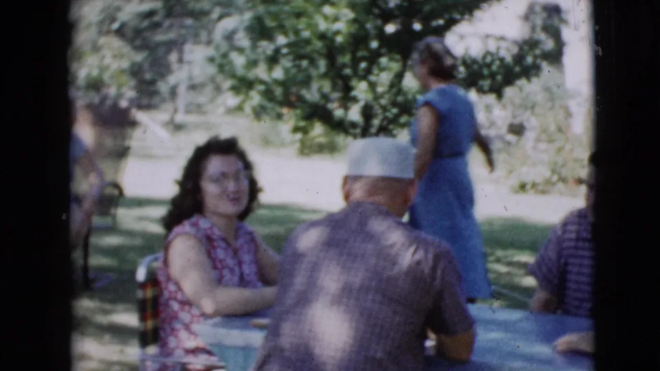 1968 Clarksdale, Arizona home-movie frame showing adults seated at a blue outdoor table with a woman walking behind them.