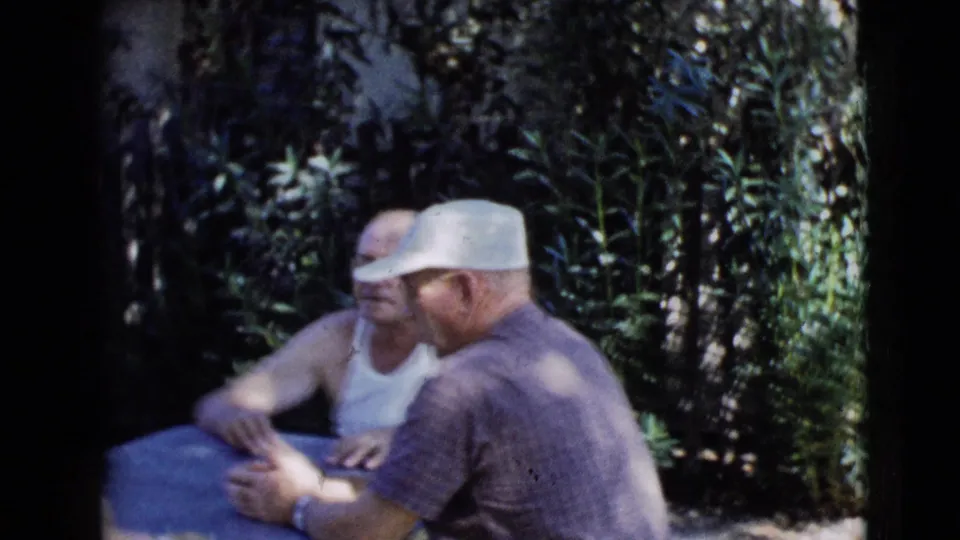 1968 Clarksdale, Arizona frame of men seated at a blue table in a leafy outdoor setting, one wearing a white cap.