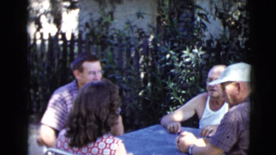 1968 Clarksdale, Arizona frame of adults around a blue outdoor table, filmed in soft 8mm color and shade.