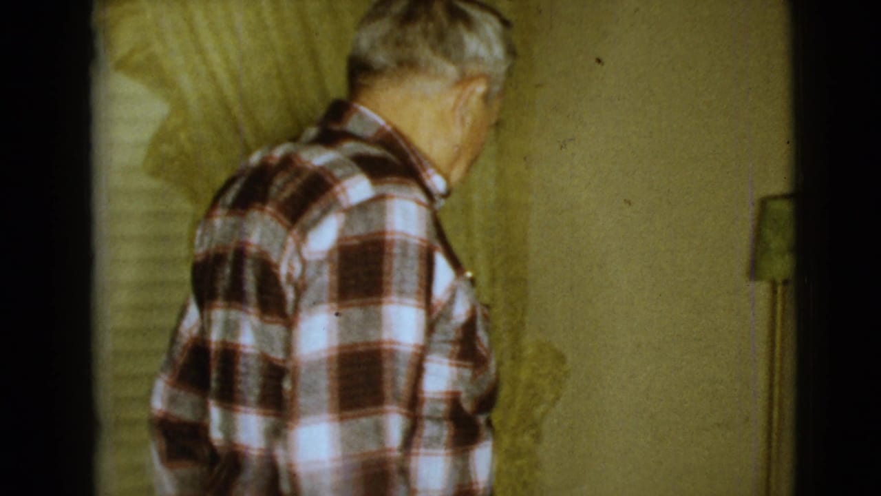 Elderly man in a plaid shirt turned away inside a dim room with curtains.