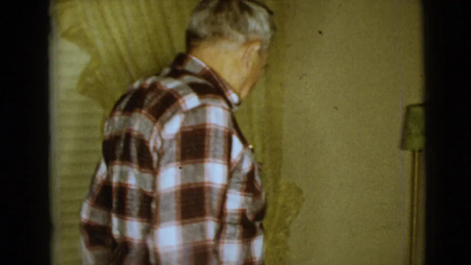 In a 1968 Clarksdale, Arizona home interior, an elderly man in a plaid shirt stands turned away near curtains and a yellow wall.