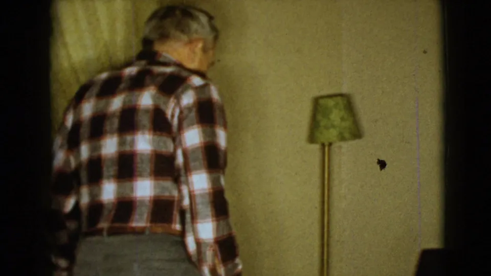 The elderly man walks away from the camera inside a 1968 Clarksdale, Arizona room with a green-shaded floor lamp and yellow wall.