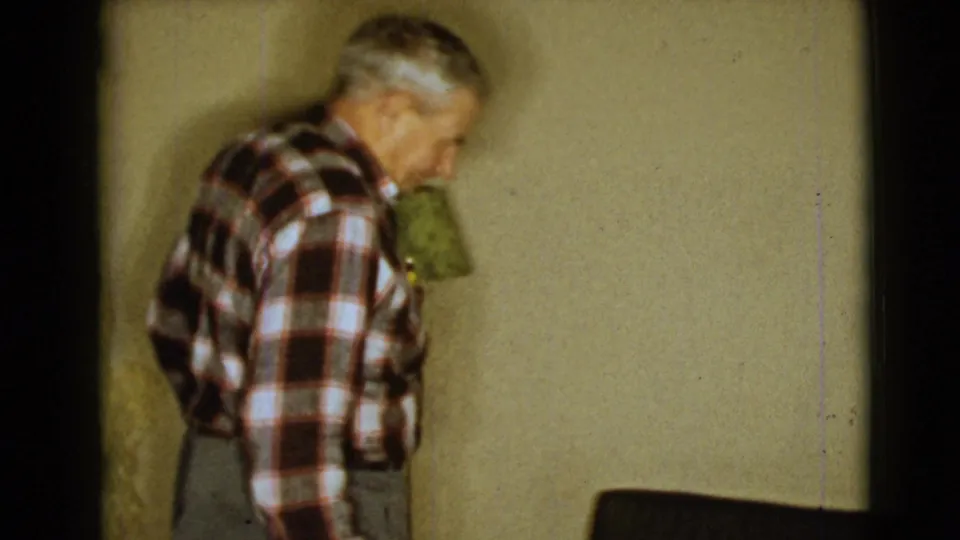 In the private Arizona home interior, the elderly man lowers his head as he walks near a green-shaded lamp and dark chair or couch edge.
