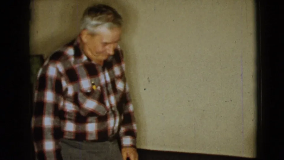 The man in a red, white, and brown plaid shirt looks downward in the 1968 Clarksdale, Arizona home-movie interior.
