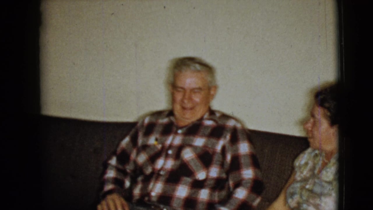 Elderly man in a red, black, and white plaid shirt smiles beside an elderly woman on a dark couch in 1968 home-movie footage.