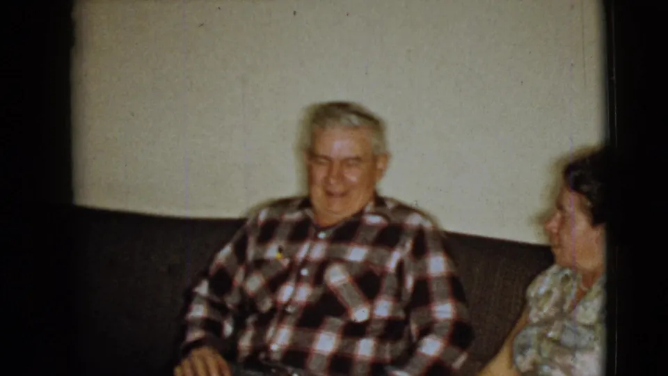 Elderly man in a red, black, and white plaid shirt smiles beside an elderly woman on a dark couch in 1968 home-movie footage.