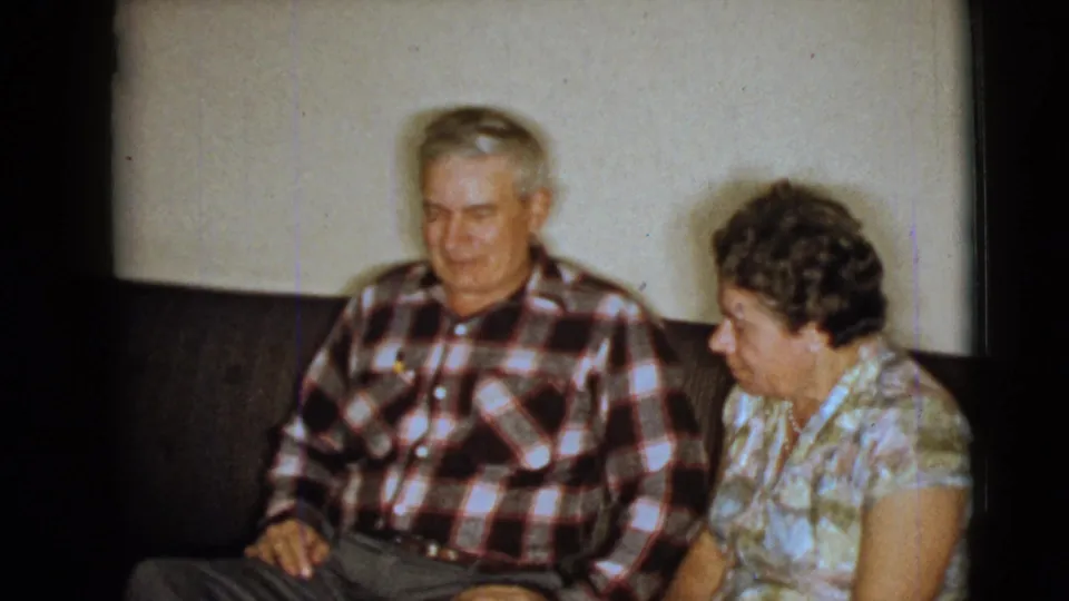 Elderly couple sit close together on a dark couch in a private interior, captured on 1968 8mm home-movie footage.