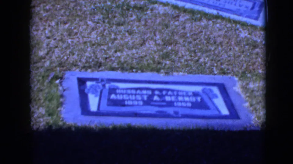 Home-movie view of a blue-tinted cemetery marker in grass, partially soft from handheld 8mm filming.