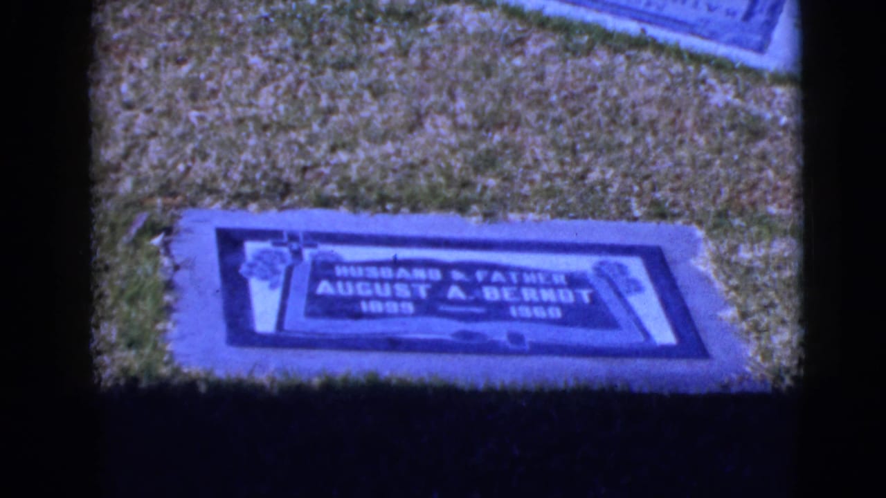 1968 8mm home-movie frame of a flat grave marker in a grassy cemetery in Clarksdale, Arizona, with cool blue film tint.