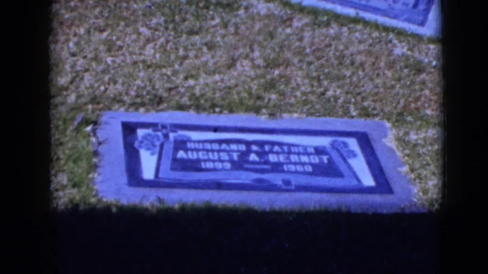 Handheld 8mm view of a flat cemetery marker and surrounding grass, with the marker text partly legible in the frame.